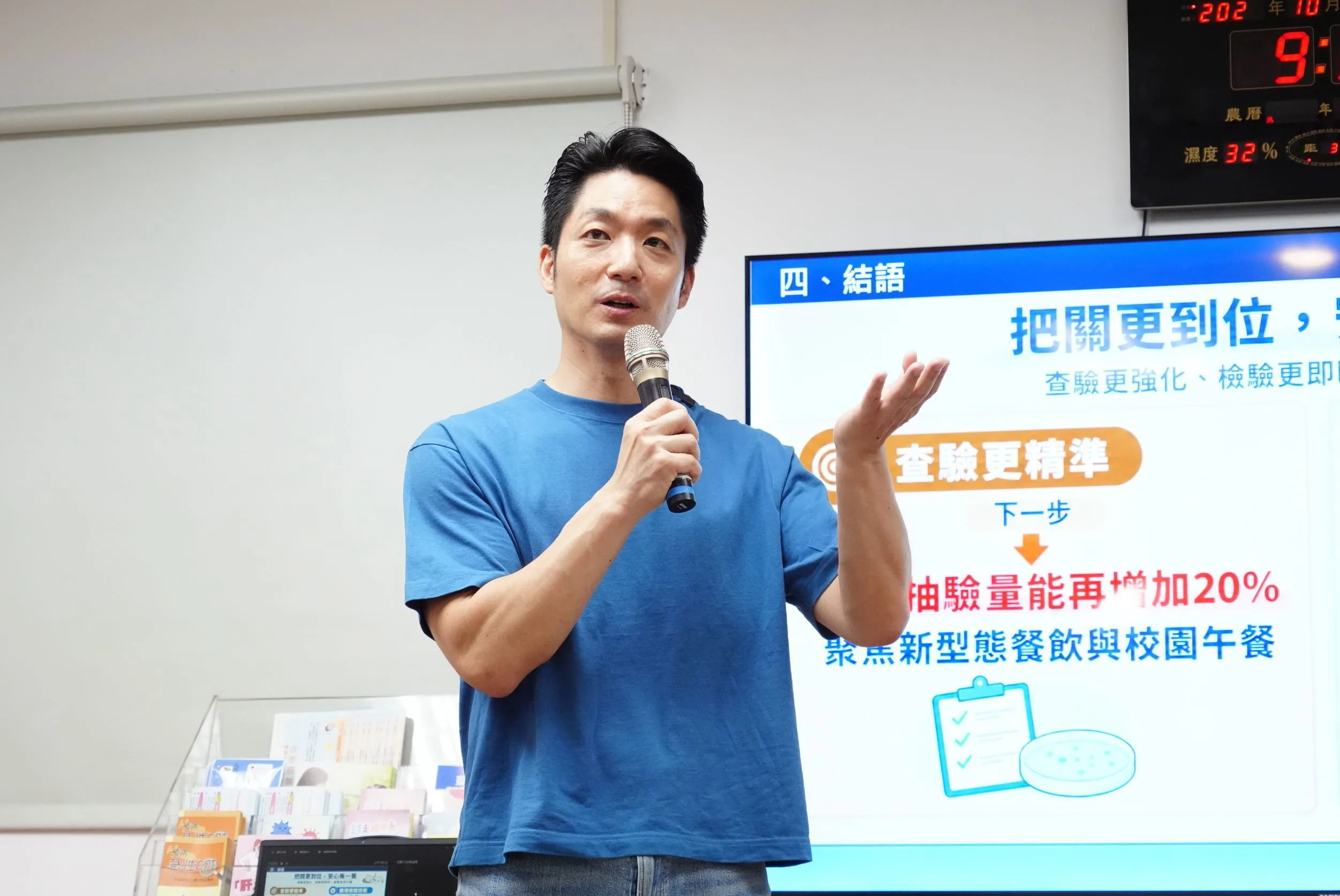 蔣萬安穿藍色上衣，手持麥克風在食安檢驗簡報前發言